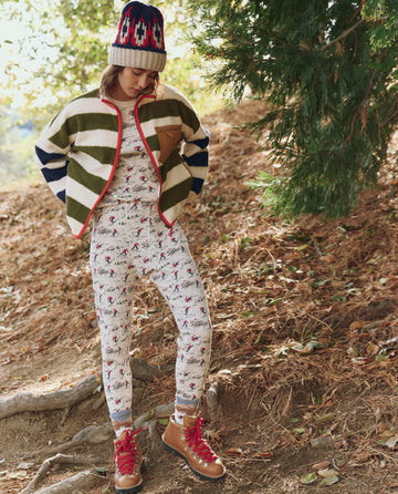 A person in THE GREAT. F25 TGO Plush Terrain Full-Zip in Mixed Heritage Stripe stands outdoors on leafy, wooded ground.