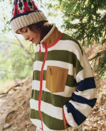 Outdoors, a person stands near trees looking downward, wearing THE GREAT. F25 TGO’s Plush Terrain Full-Zip in Mixed Heritage Stripe with a tan chest pocket and a knitted hat.