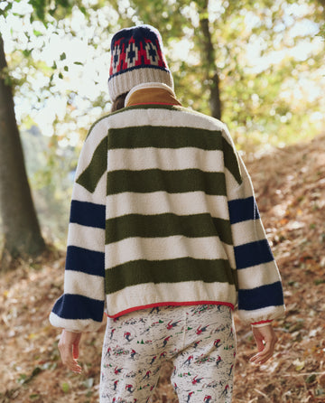 Wearing THE GREAT. F25 TGO's The Plush Terrain Full-Zip in Mixed Heritage Stripe, a person strolls through autumn woods, leaves scattered beneath their feet.