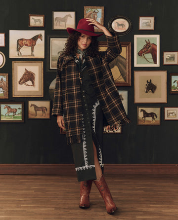 A woman wearing THE GREAT. F26 STETSON The Fringe Coat in Carmel and Black Frontier Plaid stands atop a wooden floor, framed by horse-themed artwork, red hat, and cowboy boots—perfectly channeling classic American heritage style.
