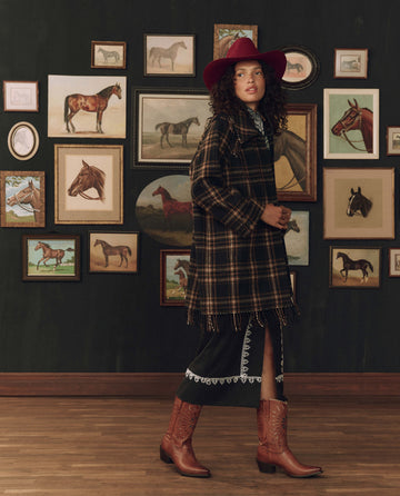 Wearing THE GREAT. F26 STETSON’s The Fringe Coat in Carmel and Black Frontier Plaid, a woman stands before a wall adorned with framed horse paintings, her black dress, red hat, and cowboy boots evoking the spirit of American heritage.