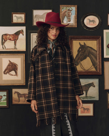 A woman in a red hat and THE GREAT. F26 STETSON “The Fringe Coat” in Carmel and Black Frontier Plaid stands before a wall adorned with framed horse paintings and drawings, evoking a sense of classic American heritage.