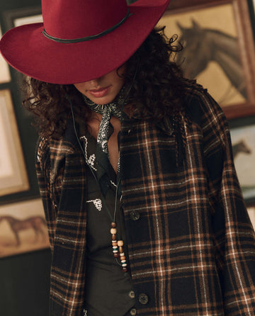 A person wearing the Fringe Coat in caramel and black frontier plaid by THE GREAT. F26 STETSON, along with a red cowboy hat and bandana, stands indoors with framed horse art in the background, capturing the spirit of American heritage.