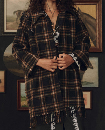 A person wearing THE GREAT. F26 STETSON’s The Fringe Coat in Carmel and Black Frontier Plaid stands in front of a wall adorned with framed paintings of horses and landscapes, evoking a sense of American heritage.