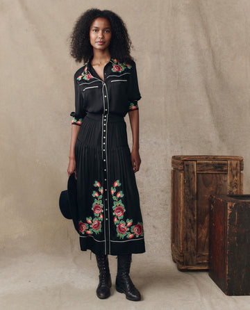 A woman stands indoors wearing THE GREAT. H25 COLLECTION's The Heirloom Rose Highland Skirt in black, paired with black boots and holding a black hat. Two wooden crates sit on the floor beside her.