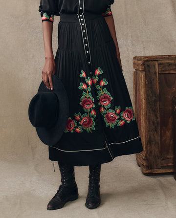 A person wearing THE GREAT. H25 COLLECTION's The Heirloom Rose Highland Skirt in black stands next to a wooden crate, paired with black boots and holding a black wide-brimmed hat against a neutral background.