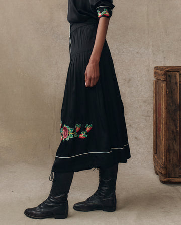 A person stands sideways against a neutral background wearing THE GREAT. H25 COLLECTION’s The Heirloom Rose Highland Skirt in black with floral embroidery near the hem, paired with black lace-up boots.