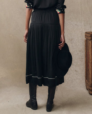 A person stands indoors facing away, wearing THE GREAT. H25 COLLECTION's The Heirloom Rose Highland Skirt in black, paired with tall black boots and holding a black hat by their side.