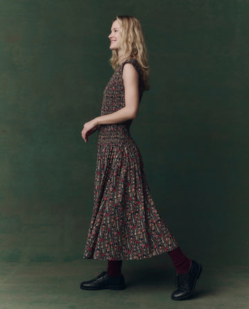 Wearing THE GREAT. F25 D2's The Paramour Dress in Garland Print, a person stands in profile against a green background, paired with dark shoes and socks.
