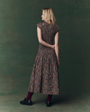 A woman stands facing away in THE GREAT. F25 D2's The Paramour Dress -- Garland Print, an ankle-length dress with a dropped waist, styled with maroon socks and black shoes against a dark green background.