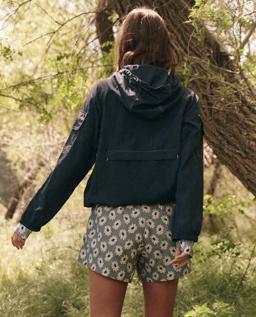 A person, clad in The Trailhead Pullover by THE GREAT. SP25 TGO--Navy and floral shorts, stands at a trailhead amidst trees and lush greenery.