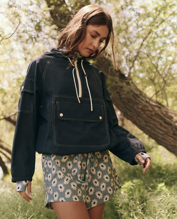 A woman in The Trailhead Pullover by THE GREAT. SP25 TGO, navy, stands at the trailhead in floral shorts, gazing downward near the trees.