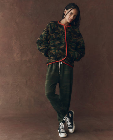 A person wearing THE GREAT. F25 D1 The Reversible Plush Bomber in Camo & Hot Red stands against a brown background, paired with green camo pants and black Converse sneakers.