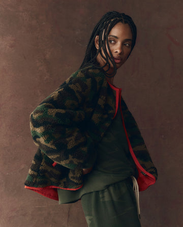 A person with braided hair models THE GREAT. F25 D1 "The Reversible Plush Bomber – Camo & Hot Red," wearing it over green clothing against a brown textured background.