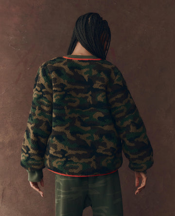 Person with braided hair faces away in THE GREAT. F25 D1 The Reversible Plush Bomber—Camo & Hot Red—paired with green pants, against a plain brown background.