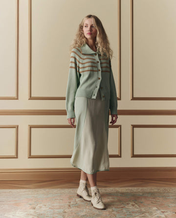 A woman stands indoors in a light green skirt, The Scholar Cardigan W/ Crochet Stripes in Seafoam by THE GREAT. SP26 D2, and beige lace-up boots against a cream wall with decorative molding.