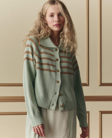 A woman with long wavy blonde hair wears THE GREAT. SP26 D2 The Scholar Cardigan W/ Crochet Stripes in Seafoam over a light dress, standing before a beige paneled wall.