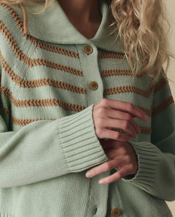 A person with wavy blonde hair wears THE GREAT. SP26 D2 The Scholar Cardigan W/ Crochet Stripes in Seafoam, featuring brown crochet stripes and buttons, showing their hands and upper torso.