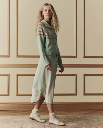 A woman stands indoors in front of a paneled wall, wearing THE GREAT. SP26 D2 The Scholar Cardigan W/ Crochet Stripes in Seafoam, paired with a light green skirt and beige lace-up boots.
