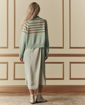 Facing a beige paneled wall, a person with long, wavy hair wears THE GREAT.'s The Scholar Cardigan W/ Crochet Stripes in Seafoam (SP26 D2), paired with a light green skirt and socks with sandals.