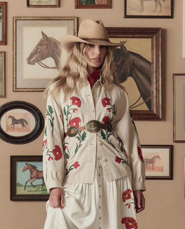 A woman wears THE GREAT. STETSON SP26 The Western Embroidered Commodore Jacket in cream and a cowboy hat, standing before a wall adorned with framed horse paintings and photos.