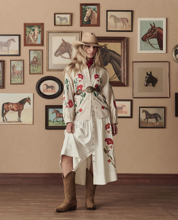 A woman in a cowboy hat and embroidered dress wears THE GREAT. STETSON SP26 The Cowboy Boot (Suede) in tan, standing indoors against a wall with framed horse pictures.