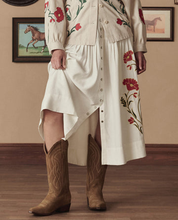 A person wearing a cream skirt and floral embroidered top, paired with THE GREAT. STETSON SP26 The Cowboy Boot (Suede) in Tan, stands indoors beside framed horse paintings.