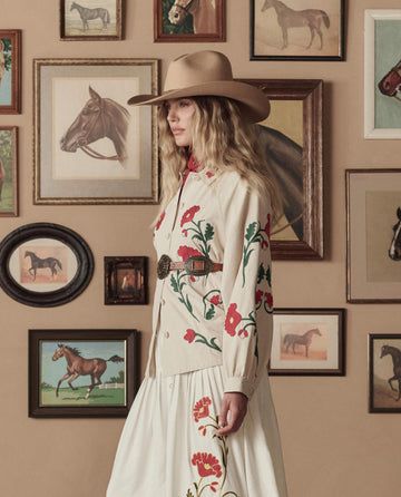 A woman in a wide-brimmed hat and THE GREAT. STETSON SP26's Western Embroidered Commodore Jacket in Cream stands before a wall adorned with framed horse artwork.