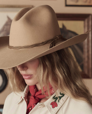 A person with long, wavy blonde hair wears THE GREAT. STETSON SP26 The Gus 6X Hat in Butterscotch, a red bandana, and a cream floral-embroidered shirt, standing before a wall decorated with framed pictures.