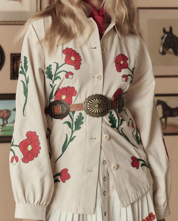 A person wears THE GREAT. STETSON SP26’s Western Embroidered Commodore Jacket in Cream, styled with a decorative belt and pleated skirt, standing before framed horse art for a classic Western-inspired look.