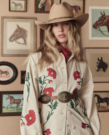 A woman in a cowboy hat and THE GREAT. STETSON SP26 Western Embroidered Commodore Jacket—Cream stands before a wall adorned with framed horse pictures.
