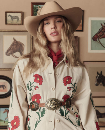 A woman in THE GREAT. STETSON SP26 The Gus 6X Hat – Butterscotch and a red-flowered embroidered shirt stands before a wall decorated with framed horse art.