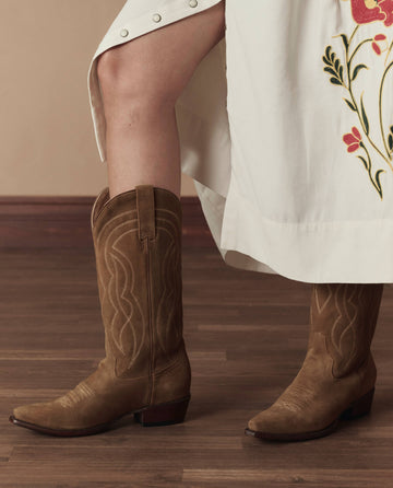 A person stands on a wooden floor in THE GREAT. STETSON SP26 The Cowboy Boot (Suede) in tan, paired with a cream floral-embroidered skirt—highlighting classic western style with a modern touch.