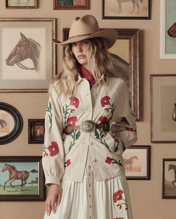 A woman wearing THE GREAT. STETSON SP26’s Tooled Leather Concho Belt in Cognac with Brass stands before a wall adorned with framed horse paintings and illustrations, styled in a beige hat and an embroidered floral outfit.