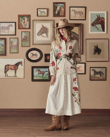 A woman in a cowboy hat and THE GREAT. STETSON SP26 Western Embroidered Commodore Jacket in cream stands indoors before a wall adorned with framed horse paintings, pairing her look with classic cowboy boots.