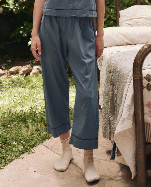 Outdoors beside a bed, only the lower body of a person is visible as they wear THE GREAT. SP25 SLEEP's The Western Easy Sleep Pant in Blue Dusk, paired with a matching top and cream-colored socks.