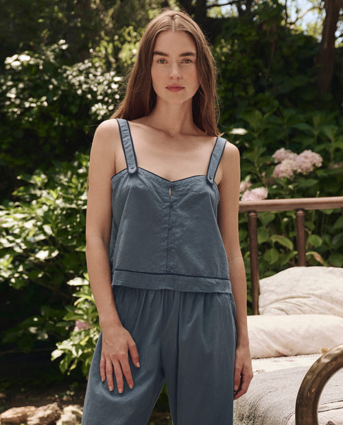 A woman with long brown hair stands outdoors in THE GREAT. SP25 SLEEP's The Western Sleep Cami in Blue Dusk, paired with matching pants, surrounded by greenery and outdoor furniture.