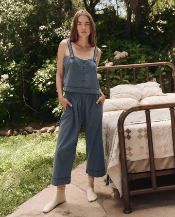 A woman stands outdoors by a vintage bed wearing THE GREAT. SP25 SLEEP's The Western Easy Sleep Pant in Blue Dusk with a blue sleeveless top and cream socks, radiating comfort among lush greenery.