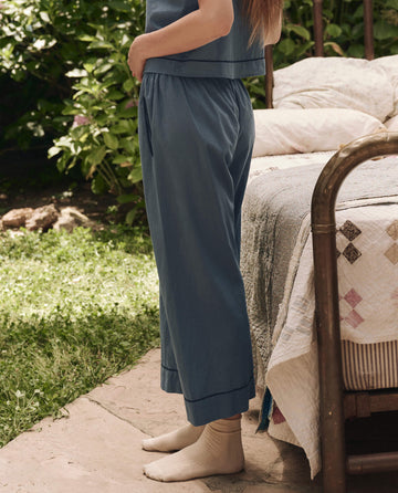 A person in The Western Easy Sleep Pant – Blue Dusk by THE GREAT. SP25 SLEEP and cream socks stands beside a bed with a quilted blanket in a sunlit garden, embodying cozy comfort wear.