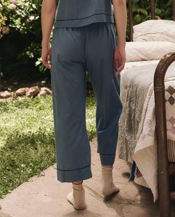 A person stands on a stone patio beside a quilted bed, wearing THE GREAT. SP25 SLEEP’s The Western Easy Sleep Pant in Blue Dusk and beige socks.