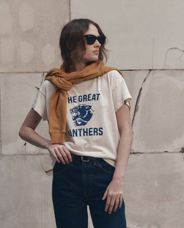 A woman wearing sunglasses, The Boxy Crew w/ Growling Panther Graphic in Washed White by THE GREAT. F26 D1, with a mustard yellow sweater draped over her shoulders, stands in front of a stone wall.