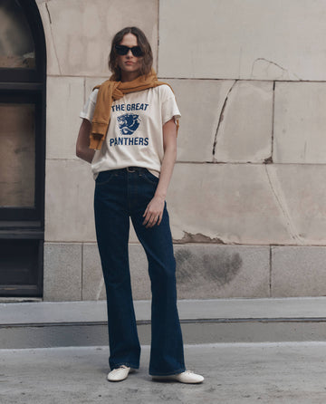 A woman in sunglasses wears THE GREAT.'s Boxy Crew w/ Growling Panther Graphic in Washed White, paired with jeans and a mustard sweater draped over her shoulders, standing against a beige stone wall.