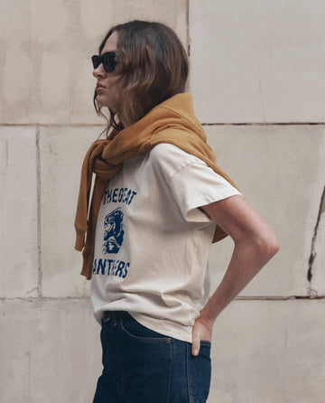 A profile shot of a person wearing sunglasses, dark jeans, and a mustard yellow sweater draped over their shoulders, styled with THE GREAT. F26 D1 The Boxy Crew w/ Growling Panther Graphic in Washed White, set against a neutral stone wall.