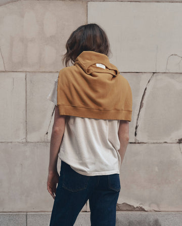 A person stands with their back to the camera, wearing THE GREAT. F26 D1’s Boxy Crew with Growling Panther Graphic in washed white, paired with dark jeans and a tan sweatshirt draped over their shoulders, in front of a stone wall.