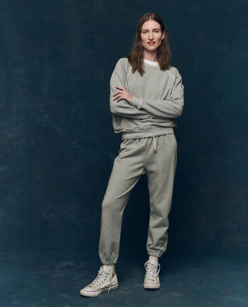 A person stands against a dark background wearing THE GREAT. CORE KNITS The Stadium Sweatpant. Solid in Varsity Grey, paired with a matching sweatshirt and white sneakers, arms crossed and smiling slightly.