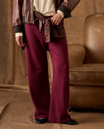 Plum-colored THE GREAT. H26 FLEECE BACK KNITS The Fleece Lap Sweatpant wide-leg fleece sweatpants with a drawstring waist, worn with a brown jacket and a plaid shirt tied at the waist.