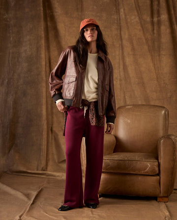 A person wearing a brown leather jacket, a cream sweatshirt, a plaid shirt tied around their waist, and plum-colored THE GREAT. H26 FLEECE BACK KNITS The Fleece Lap Sweatpant standing by a leather armchair.