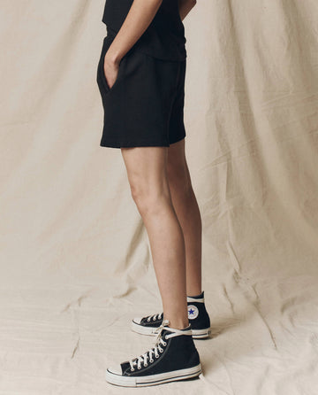 Person standing sideways with hands in pockets, wearing THE GREAT. SU25 CORE KNITS The Victory Short. Solid -- Almost Black and a black top for a comfortable fit, paired with black high-top Converse sneakers against a beige fabric backdrop.