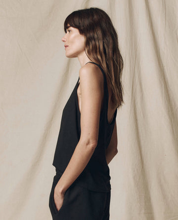 A woman with long brown hair, wearing THE GREAT. SU25 CORE KNITS The Scoop Neck Tank in Solid -- Almost Black, stands in profile against a beige fabric backdrop.