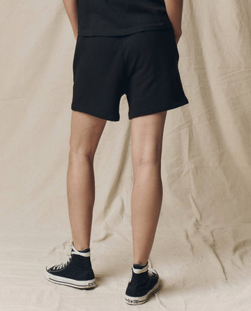 A person stands with their back to the camera wearing THE GREAT. SU25 CORE KNITS The Victory Short. Solid -- Almost Black, paired with a black shirt and high-top sneakers on a neutral fabric background, highlighting comfortable athletic wear.